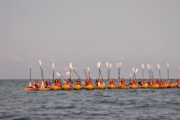 Group of kayakers in yellow kayaks at sea, paddles raised, under a pinkish evening sky.