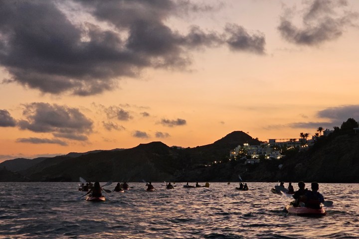 Kayakers on calm sea under cloudy orange sunset sky and silhouetted hills.