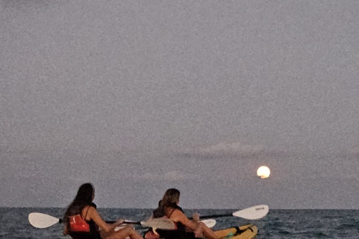 Two people kayaking on the ocean at dusk with the moon in the background.