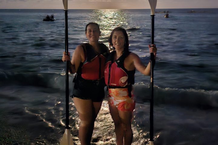 Two people with paddles on a beach at sunset with a reflective sea.