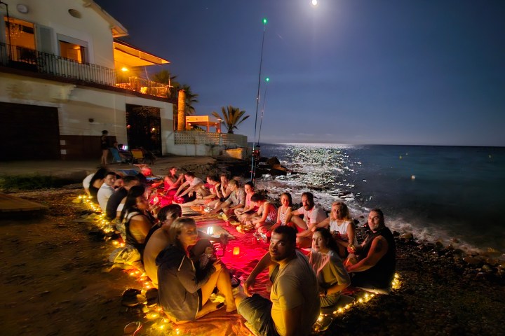 Group of people gathered on a beach at night with string lights and the moon shining.