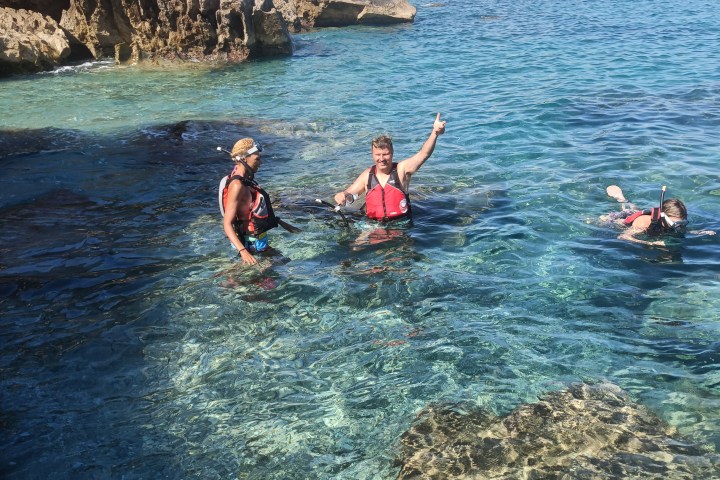 Three people snorkeling in clear blue water near rocky cliffs and coastal buildings.
