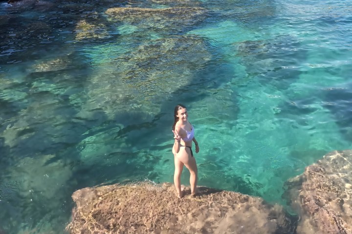 Person in swimwear stands on a rock in crystal-clear turquoise water near rocky shore.
