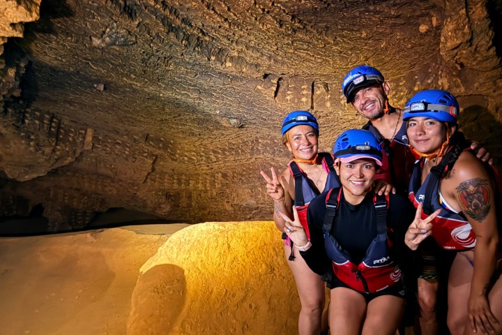 Four people in helmets posing inside a cave, smiling and holding up peace signs.