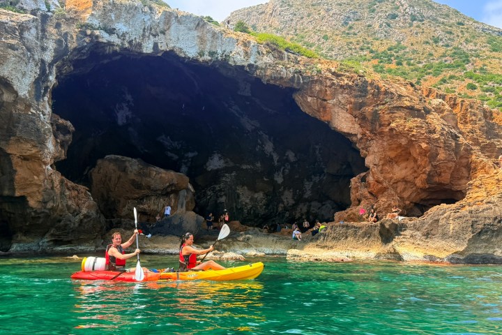 Two people kayaking near a large rocky cave opening by clear water.