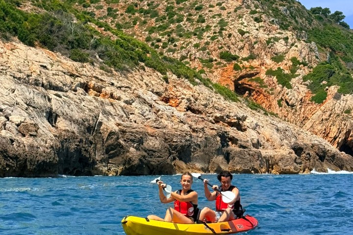 Two people kayaking on blue water near rocky shore with hills in the background.