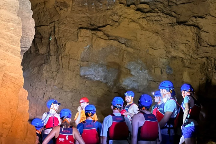 Group of people in helmets and vests inside a cave, illuminated by dim light.