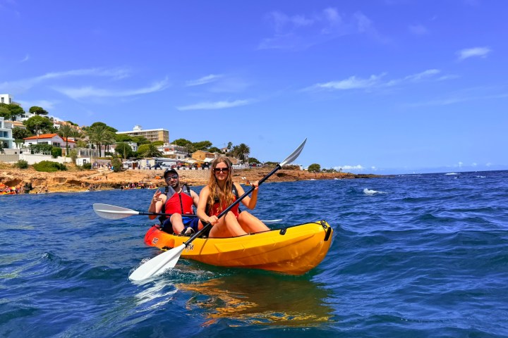 Two people kayaking on calm blue sea near rocky coastline under a clear sky.