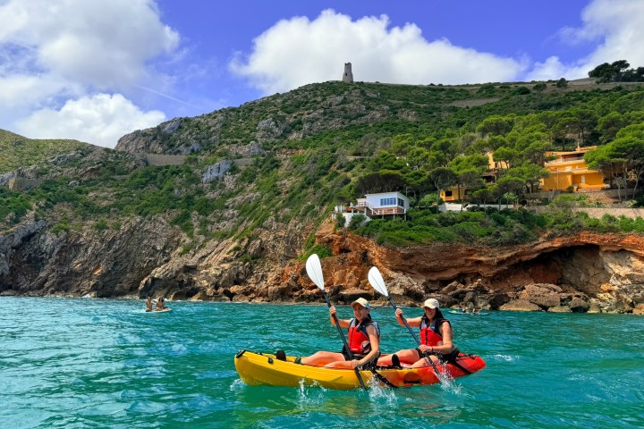 Two people kayaking on turquoise water near a rocky coastline with trees and a tower in the background.
