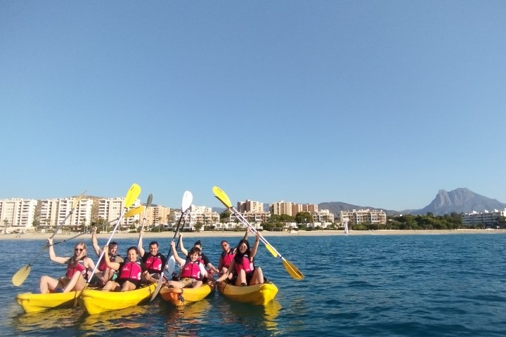 Group kayaking near a beach with buildings and mountains in the background on a clear day.