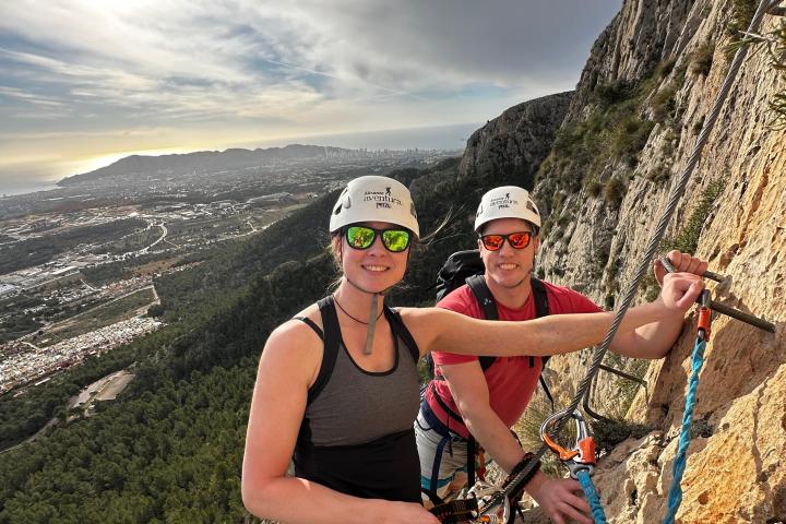 Two climbers with helmets and gear scaling a rocky cliff with a scenic view in the background.