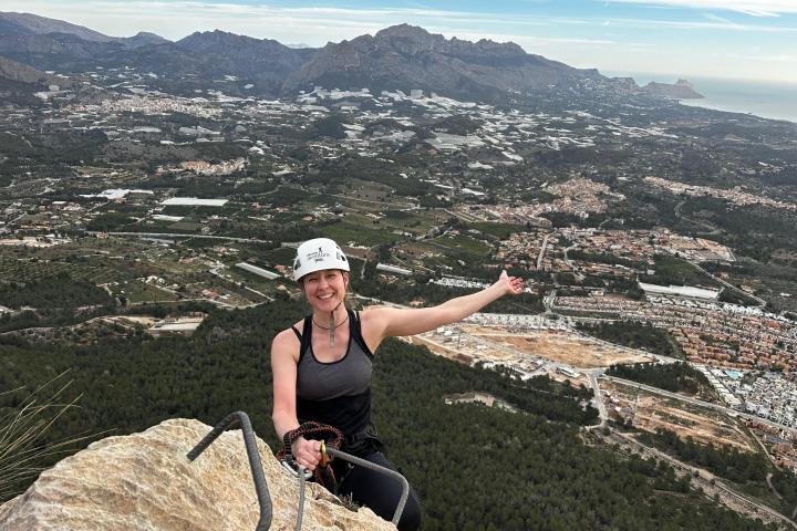 Person smiling atop a mountain with a scenic view of a valley and distant mountains.
