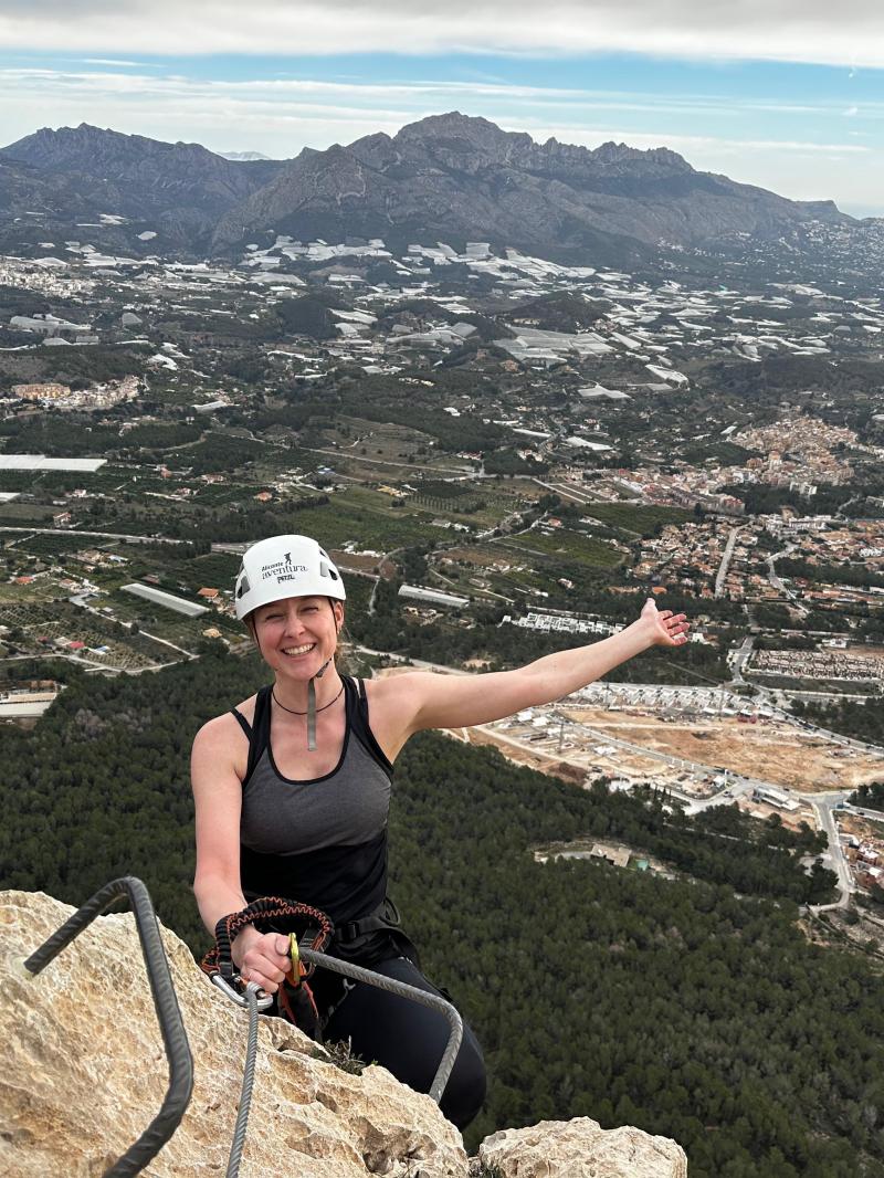 Person smiling atop a mountain with a scenic view of a valley and distant mountains.