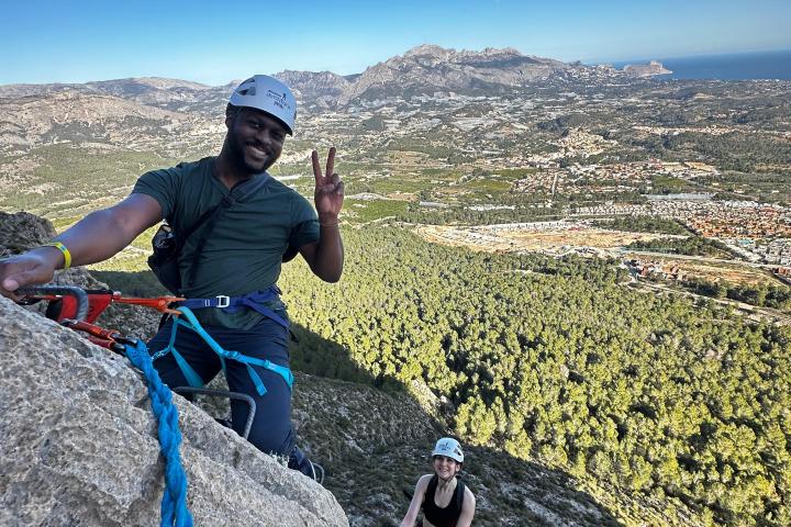 Two climbers with helmets on a steep rock face, overlooking a scenic landscape and clear blue sky.