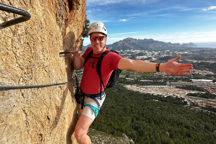 Person in a helmet and harness climbing a rocky wall with a scenic landscape below.