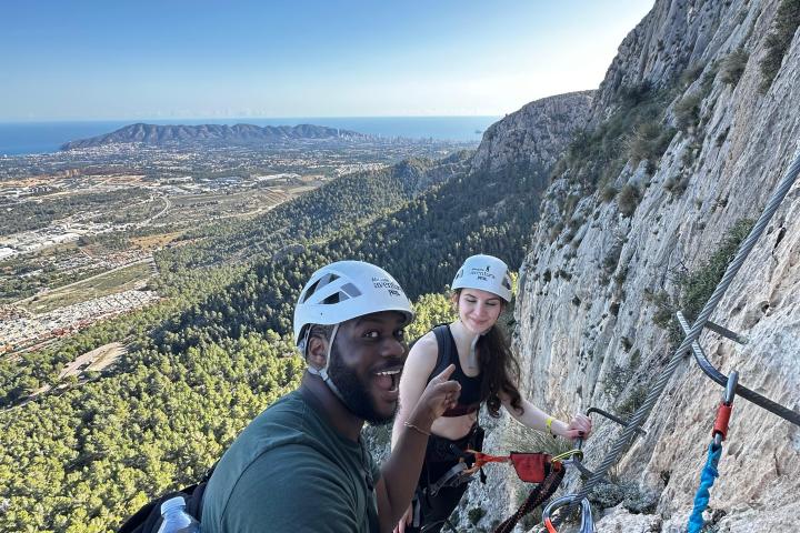 Two climbers with helmets ascend a rocky cliff with a scenic coastal view in the background.