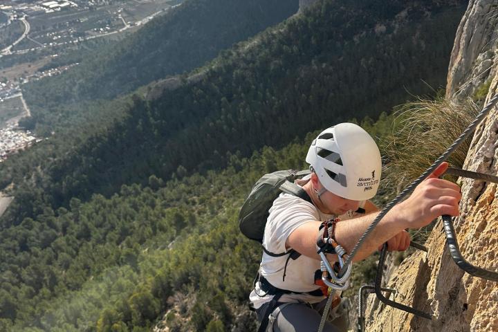 Person climbing a rocky mountain with safety gear overlooking city and sea.