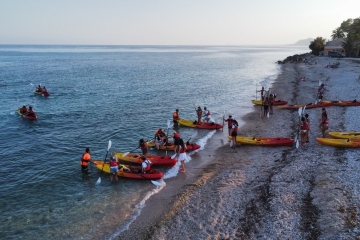 People in life jackets kayaking near a pebbled beach at sunset.