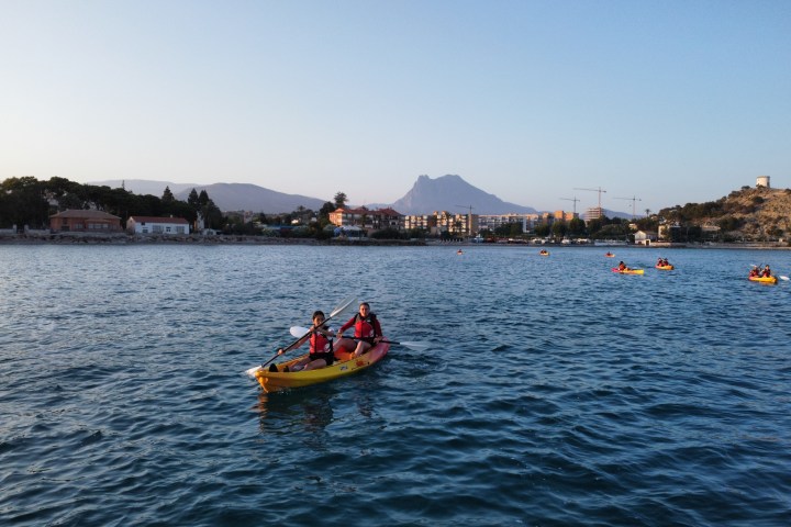 People kayaking on a calm sea with a town and mountains in the background.