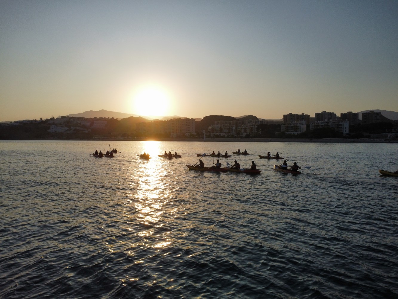 Group kayaking on a calm sea at sunrise with cityscape in background.