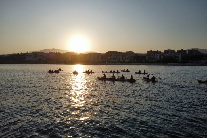 Group kayaking on a calm sea at sunrise with cityscape in background.