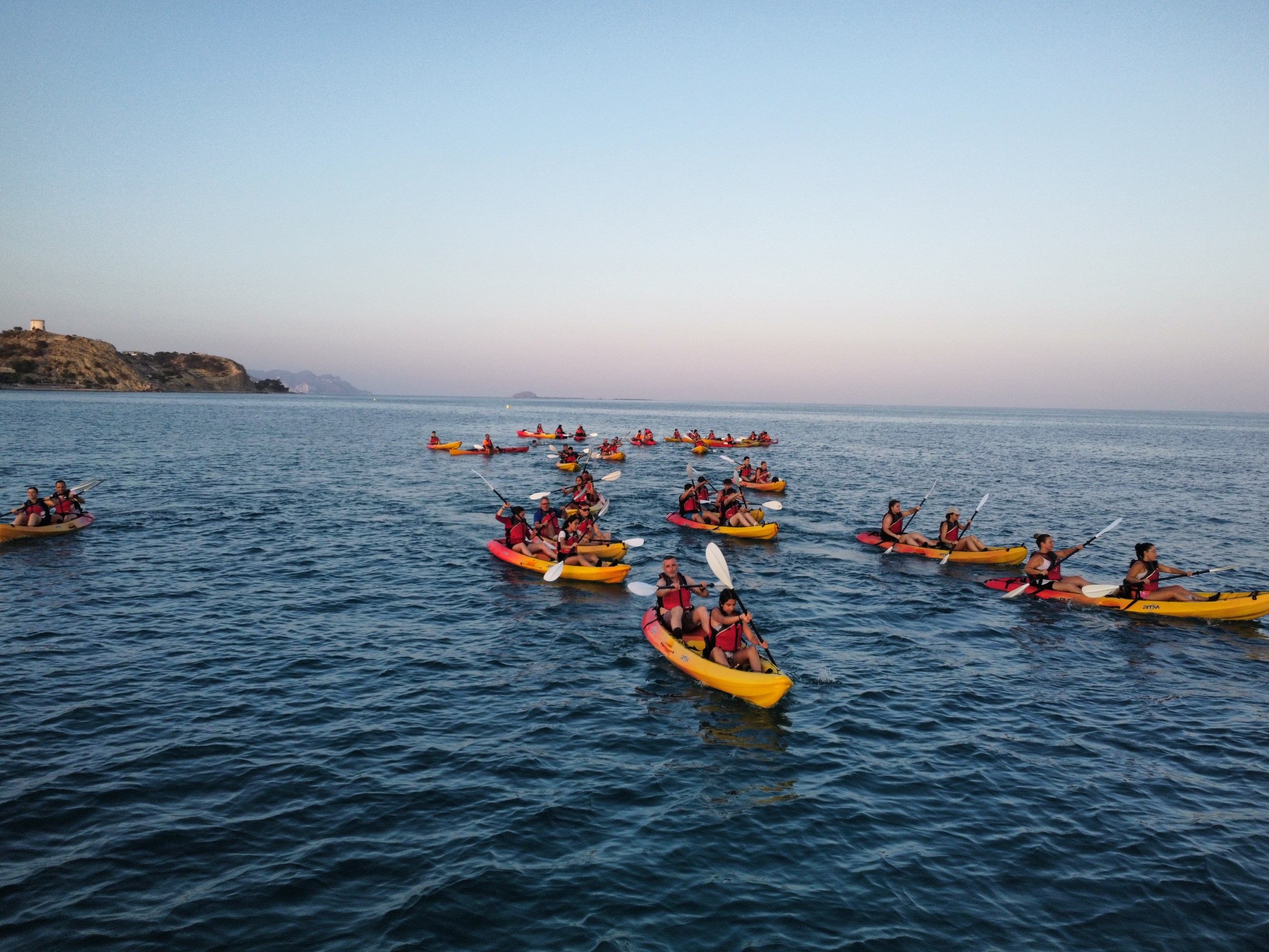 Group of people kayaking in the ocean near a rocky coastline during sunset.