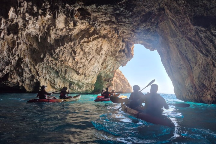 Kayakers paddling through a sunlit cave opening into a bright blue sea.