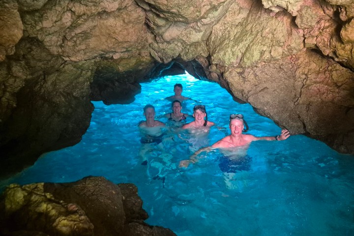 Five people swimming in a rocky cave with clear blue water.