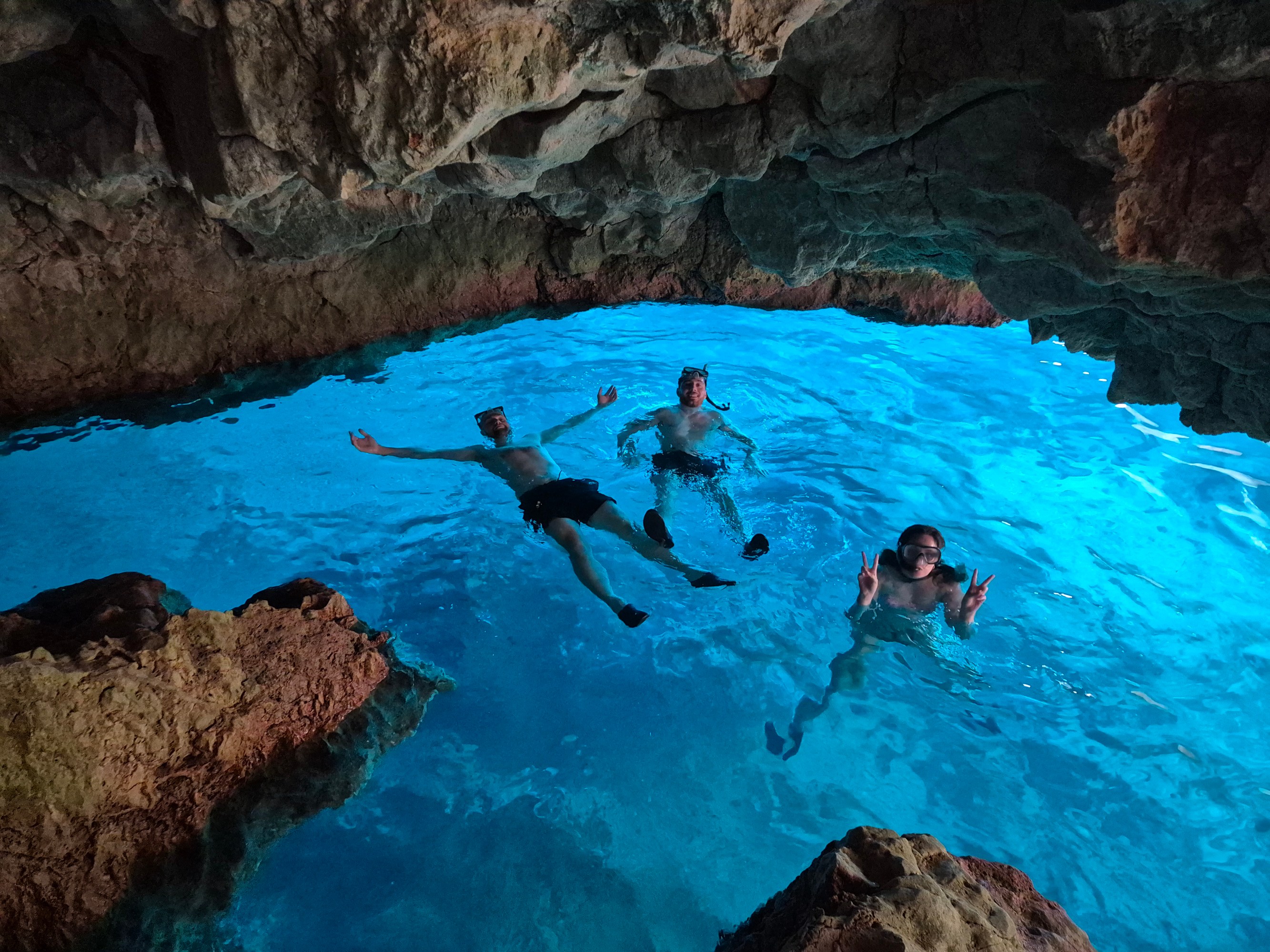Three people snorkeling in a brightly lit blue water cave with rocky ceiling.