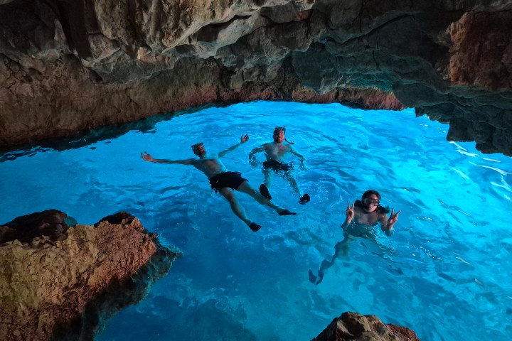 Three people snorkeling in a brightly lit blue water cave with rocky ceiling.