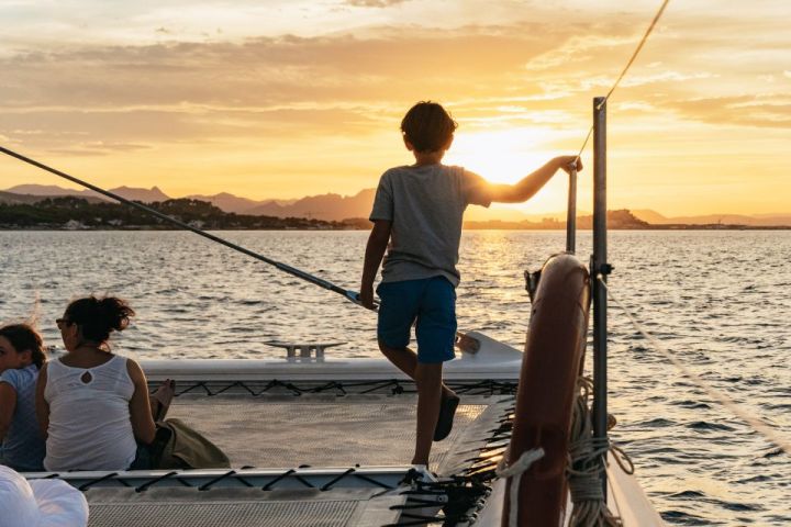 Boy standing on boat deck at sunset, with people sitting nearby.