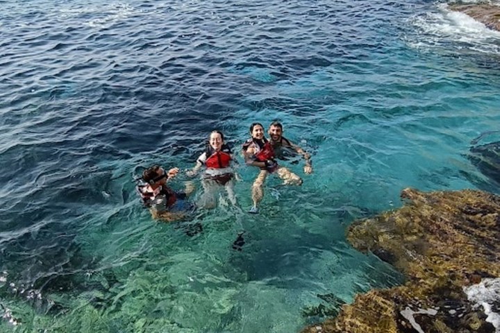 Four people in life jackets swimming near a rocky coastline in clear blue sea.