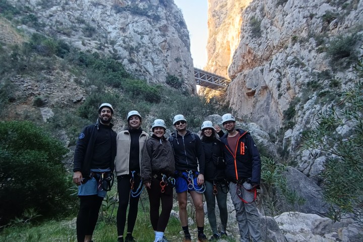 Group of six hikers with helmets in a rocky canyon with a bridge in background.