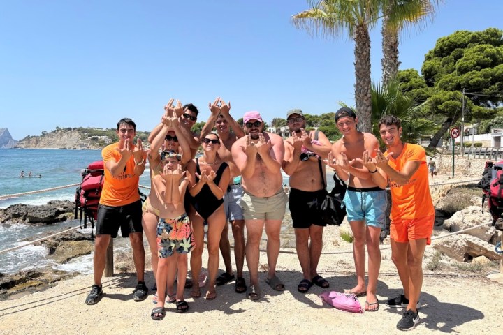 Group of people in swimwear posing on a sunny beach with palm trees and ocean in the background.