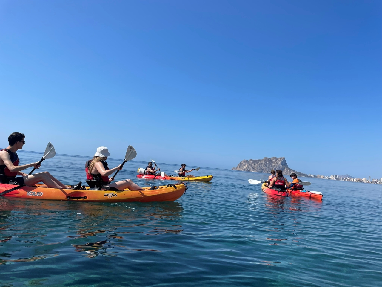 People kayaking on a calm sea with a distant rock formation under clear blue sky.