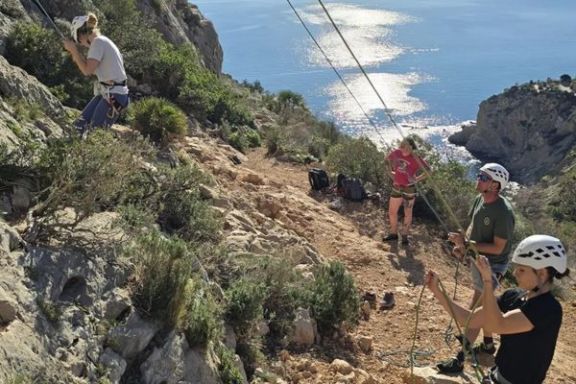 Participante practicando escalada en roca mientras otros compañeros manejan las cuerdas de seguridad junto al mar.