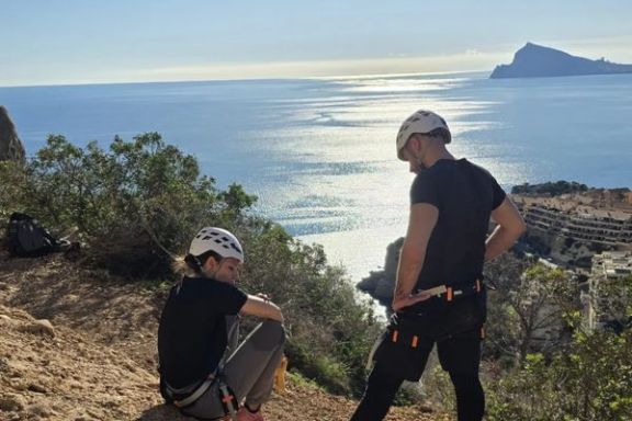 Dos participantes descansando con el equipo de escalada junto a las vistas del Mediterráneo desde el Morro de Toix.