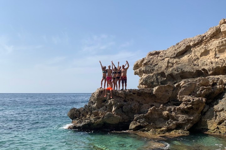 Group of people in swimwear standing on rocks by the sea, arms raised under a clear blue sky.