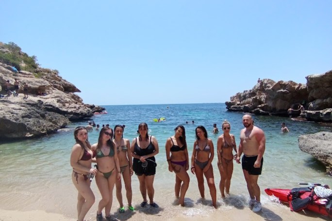 Group of people in swimwear standing on a sandy beach with a rocky shoreline and clear blue sky.