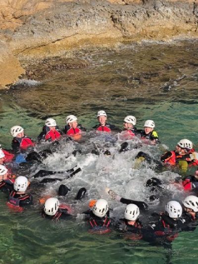 Group of people in helmets form a circle in the water near a rocky shore.
