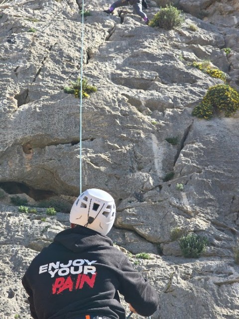 Two climbers on a rocky wall, one belaying, with 'Enjoy Your Pain' on their back.