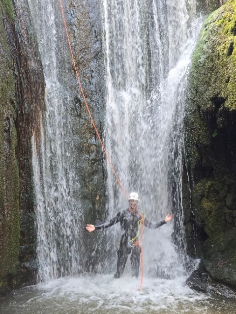 Person in helmet under waterfall with climbing gear, standing in water.