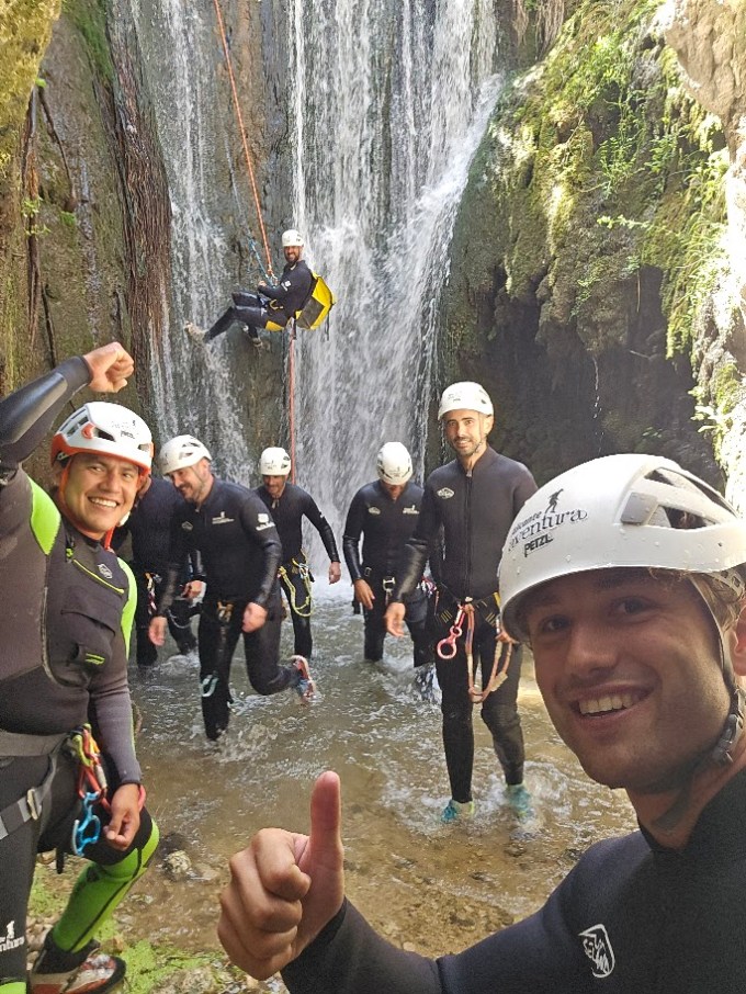 Group of people canyoning in wetsuits and helmets near a waterfall, one person rappelling.
