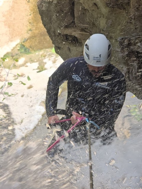 Person in helmet rappelling down a rock face with water splashing around.
