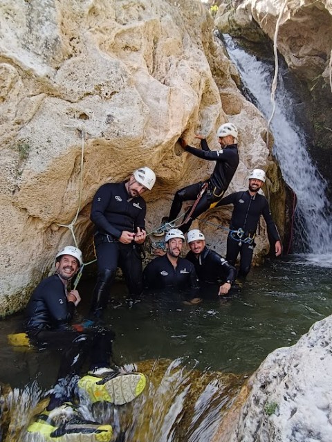 Group of people in wetsuits and helmets standing in water near a rock face and waterfall.