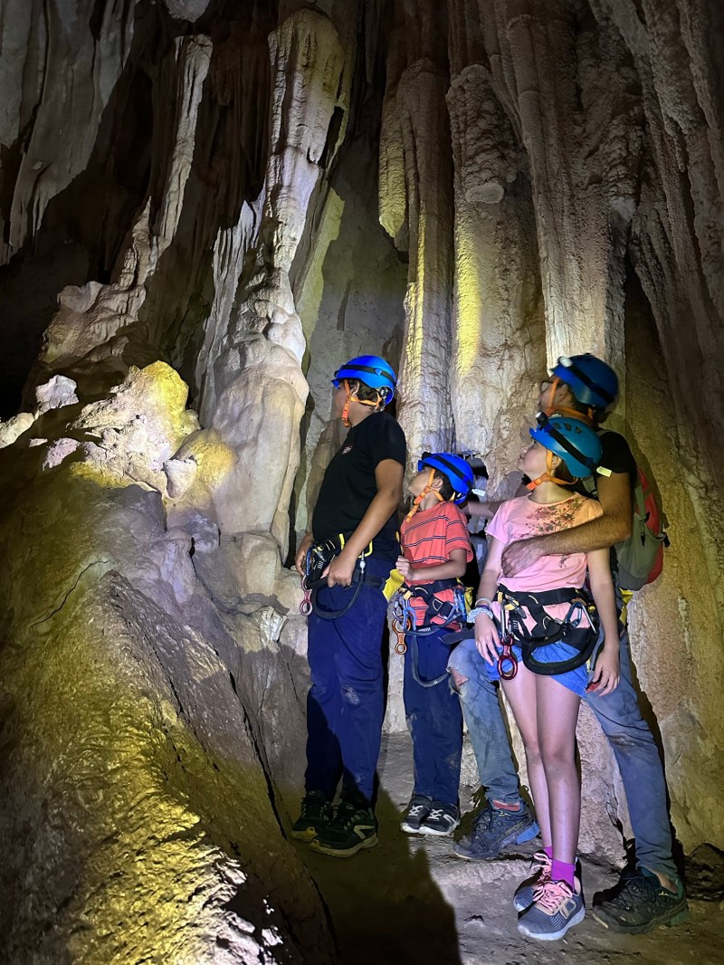 Four people with helmets and ropes exploring a cave with stalactites.