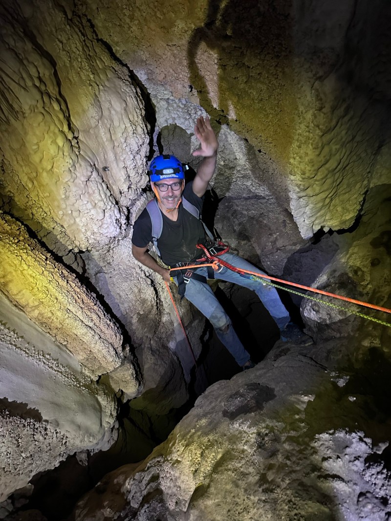 Man in a cave wearing a helmet and harness, waving while climbing with ropes.