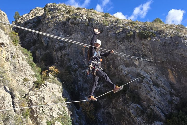 Person crossing a high rope bridge on a rocky cliff under a clear blue sky.