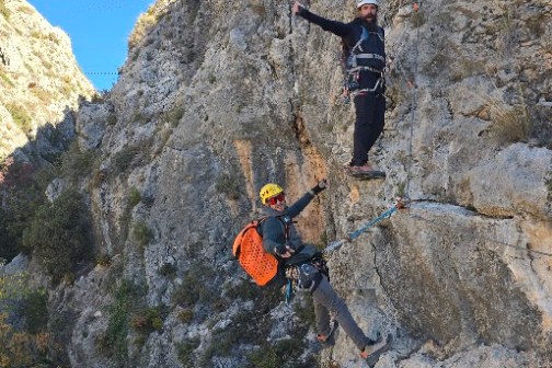 Two climbers on a steep rocky cliff with safety gear against a clear blue sky.