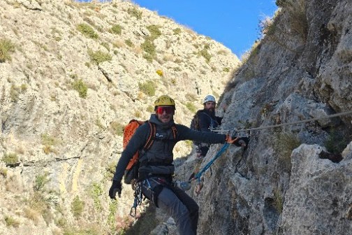 Two climbers navigating a rocky mountain path using safety ropes under a clear blue sky.